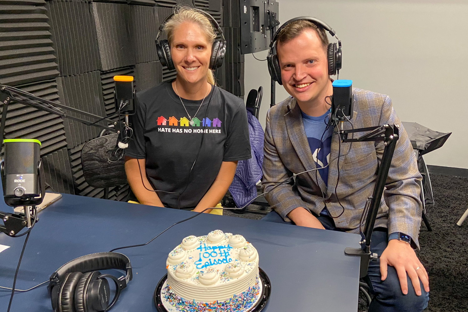 Image description: Hosts Bobbi and Alex pose in the podcast studio in front of their mics and a cake that reads 
