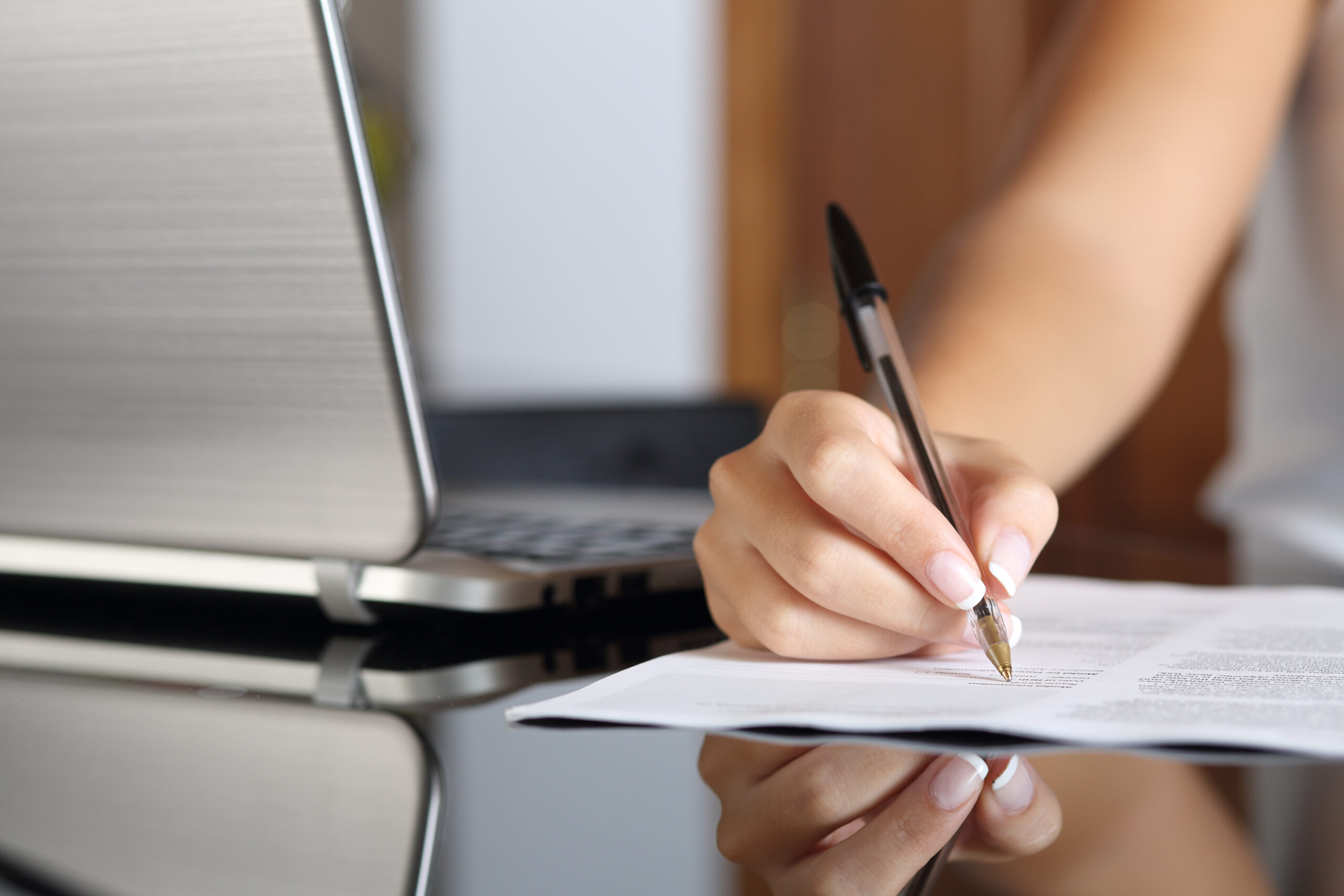 Image description: Close up of a woman hand writing a form with a laptop beside at home or office.